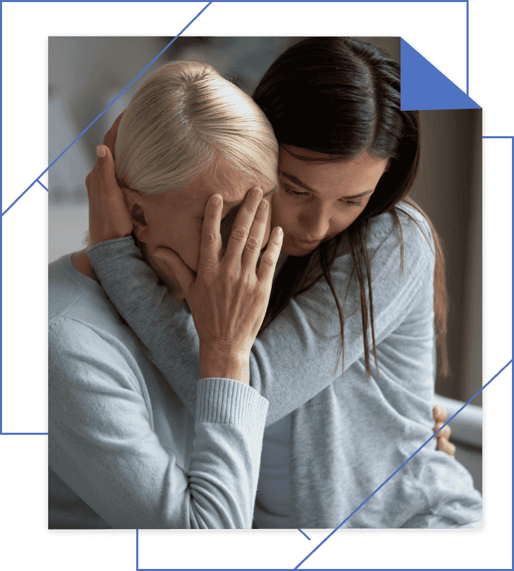 An older woman covers her face while a younger woman hugs her from the side, both in gray clothing, in a supportive gesture.