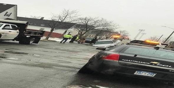 On an overcast day in Hamtramck, a police car and a tow truck assist a white van stalled near a pothole on the wet street. People stand nearby in the residential area, observing the scene.