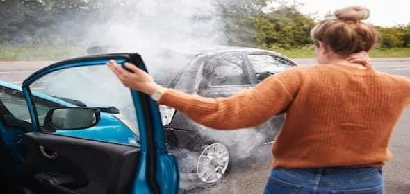 A woman stands beside an open car door, assessing a smoking vehicle on the road after what seems like one of those all-too-frequent Michigan car accidents.