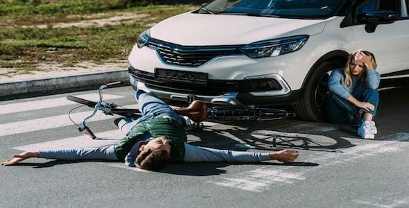 A cyclist lies on the ground at a Michigan crosswalk in front of a car, with a woman sitting nearby holding her head. Bike accidents like these highlight the importance of having car insurance.