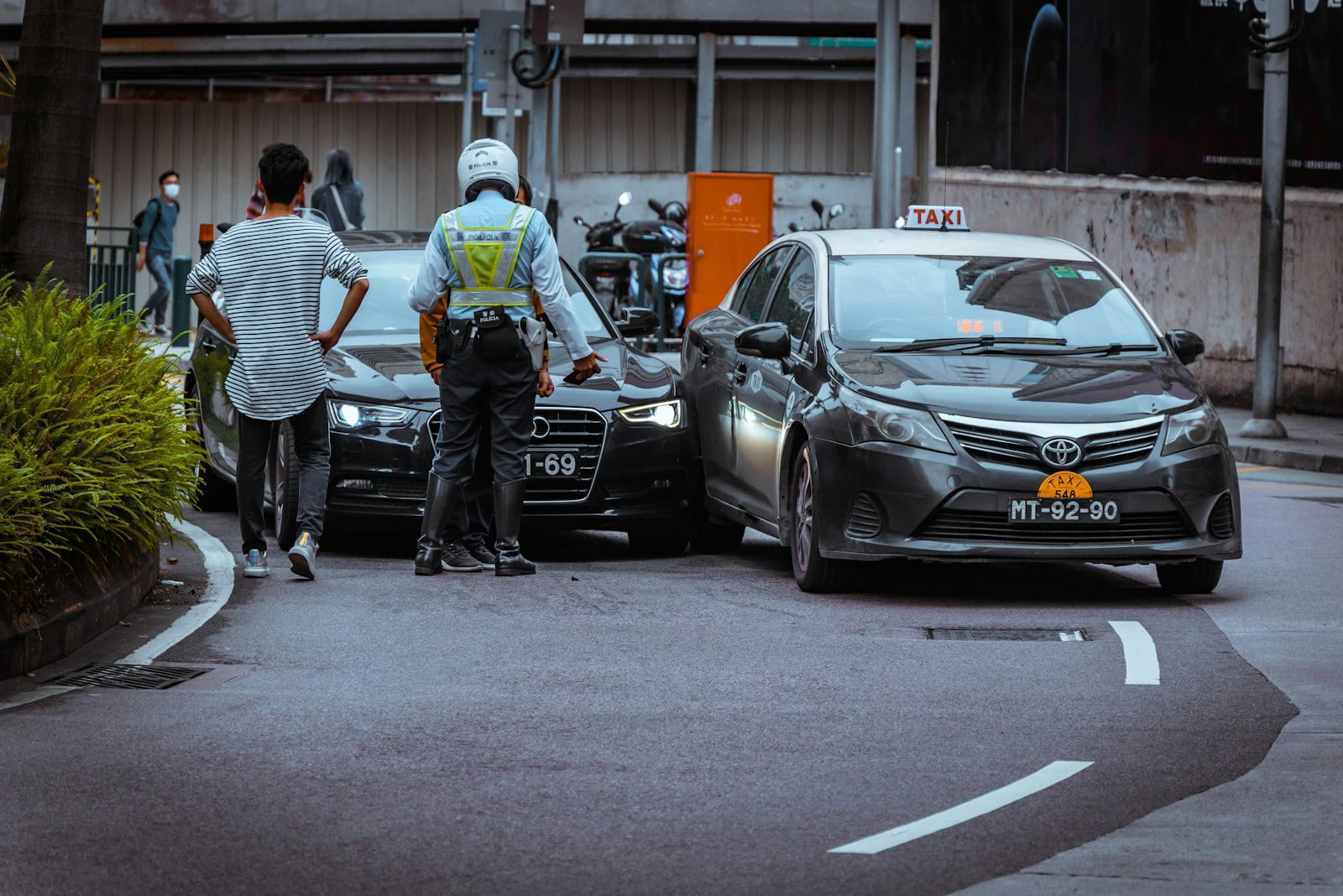 Two cars, a Mercedes and a Toyota, have been involved in a minor rear-end collision on the street. A person and a traffic officer are standing nearby.